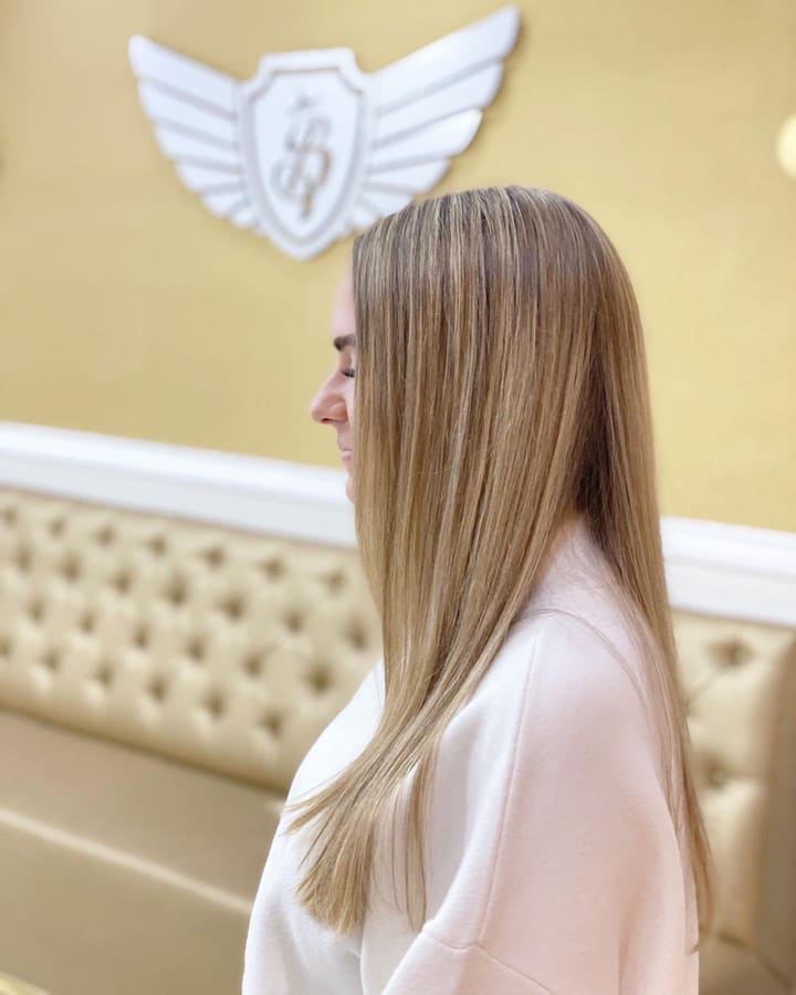 Vrouw met lang, steil blond haar voor de champagnekleurige wand met het witte vleugellogo en de crème chesterfieldbank